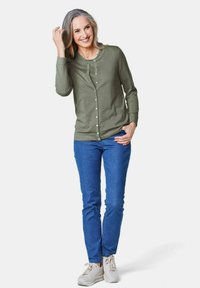 Groene geribde cardigan met knoopdetails, gedragen over blauwe spijkerbroeken. Model staat, heeft lang grijs haar en draagt witte sneakers. Eenvoudig ontwerp.