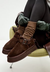 Bruine suede schoenen met een plateauzool en een gespsluiting, gedragen met geribde bruine sokken en zwarte leggings, zittend op een groene stoel.