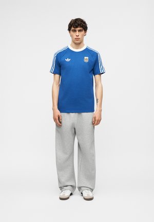 Joven de pie con una camiseta de fútbol azul de Adidas Argentina, pantalones deportivos gris claro y zapatillas blancas sobre un fondo liso.