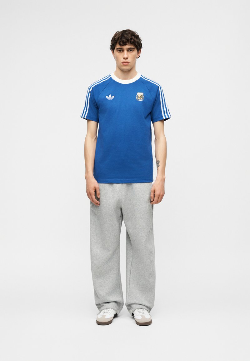 Νέος άνδρας που φορά μπλε φανέλα ποδοσφαίρου Adidas Argentina, ανοιχτό γκρι φόρμα και λευκά αθλητικά παπούτσια απέναντι σε απλό φόντο.