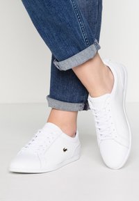 Bílé kožené tenisky s mesh detaily, kulatá špička a plochá podrážka. Zlaté logo na boku. Nošené s ohrnutými modrými džínami.