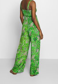 Jumpsuit floreale verde con gambe ampie, caratterizzato da un corpetto aderente e spalline regolabili. Il tessuto presenta fiori rosa e bianchi vivaci.