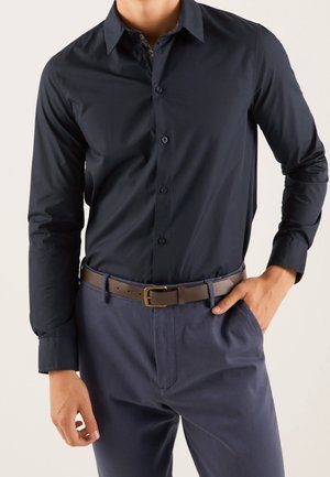 Homme portant une chemise ajustée bleu marine foncé boutonnée et rentrée dans un pantalon bleu foncé avec une ceinture en cuir marron, une main dans la poche.