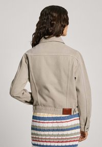 Chaqueta de mezclilla beige con un cuello clásico y puños abotonados. Cuenta con un detalle de etiqueta de cuero en la parte posterior y un corte ligeramente ajustado.