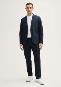 Blazer din in marin cu rever în formă de U și buzunare, asortat cu un tricou alb și pantaloni marinei asortați. Adidași albi completează ținuta.