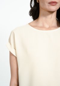 Krēmkrāsas blūze ar īsām piedurknēm un apaļu apkakli. Izceļas ar gludu tekstūru un apvītām piedurkņu malām.