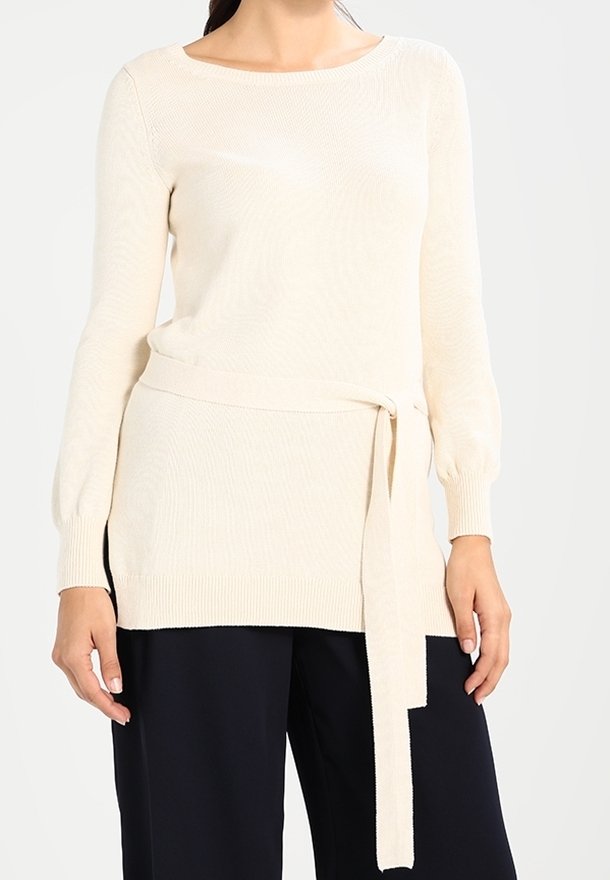 Cremfarbener Strickpullover mit langen Ärmeln, einem runden Halsausschnitt und einem gebundenen Taillendetail, kombiniert mit dunklen Hosen. Strukturierter Stofffinish.