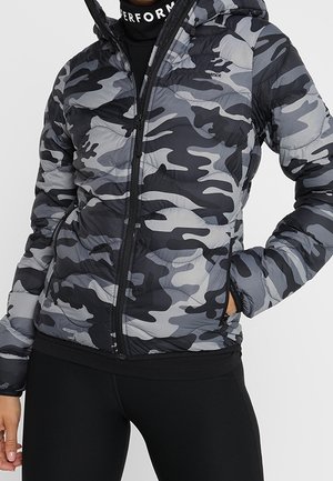 Femme portant une doudoune camouflage grise et noire avec capuche, des leggings noirs et un col roulé noir avec texte blanc.