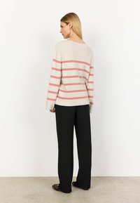 Striped sweater in beige with coral horizontal stripes, soft texture, relaxed fit, and a wide neckline; paired with black wide-leg pants and patterned shoes.