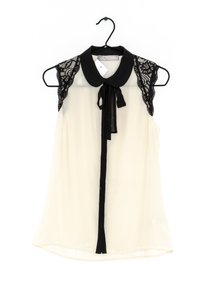 Sleeveless blouse in cream with a black lace yoke, collar, and ribbon tie. Smooth texture with a relaxed fit and rounded hem.
