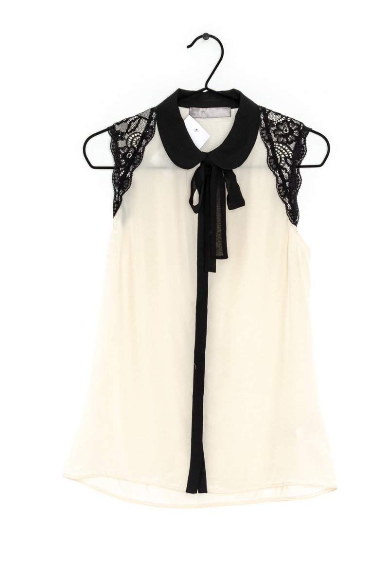 Sleeveless blouse in cream with a black lace yoke, collar, and ribbon tie. Smooth texture with a relaxed fit and rounded hem.