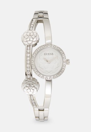 El reloj Guess plateado presenta una esfera blanca redonda con un diseño floral, rodeada por un bisel de cristales. Dos acentos florales realzan la correa de la pulsera.