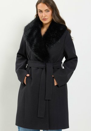 Femme portant un manteau noir ceinturé avec un large col en fourrure, les mains dans les poches, et de longs cheveux bruns, debout devant un fond clair uni.