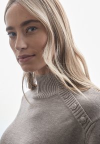 Světlé hnědé pletené svetr s vysokým žebrovaným límcem a dekorativními knoflíky na rameni. Měkká textura, přiléhavý design.