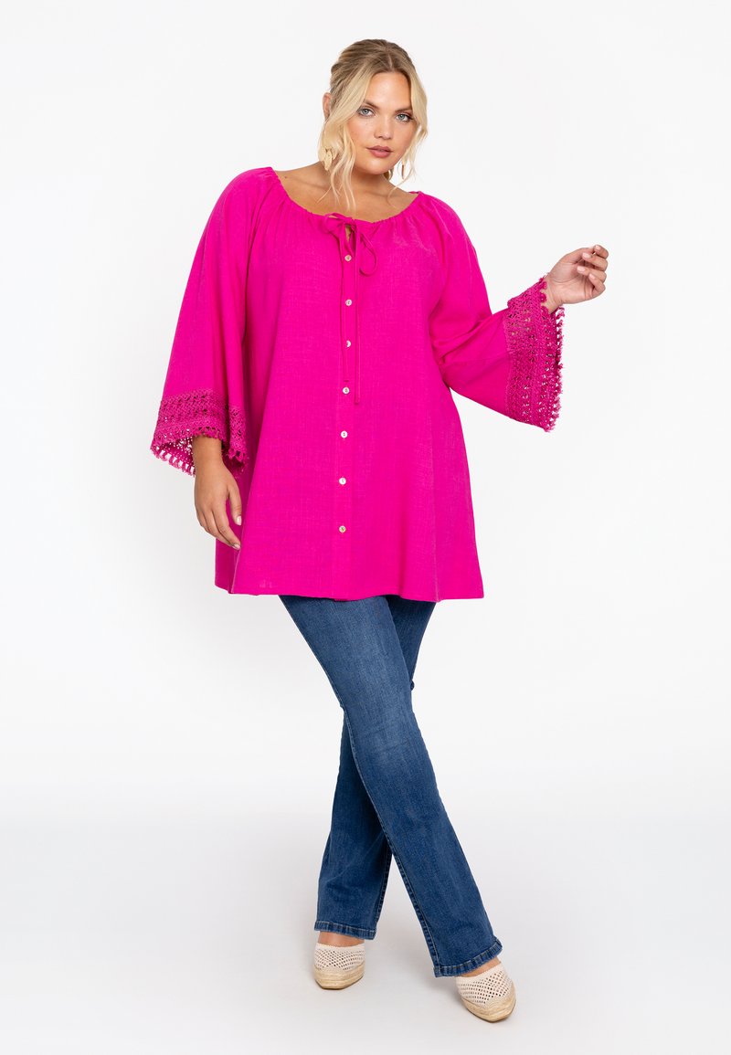 Blouse rose vif avec un col rond, détail de nœud, devant à boutons et accents en crochet sur les manches évasées, associée à un jean bleu et des chaussures compensées.