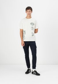 Weißes Baumwoll-T-Shirt mit grünen Grafikdrucks, kombiniert mit navyfarbenen Cargo-Hosen und schwarzen Sneakers mit weißen Streifen.