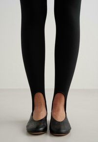 Svarta leggings med utskärningar vid anklarna, kombinerade med svarta läderbalarinas med rundad tå och låg klack, visade mot en neutral bakgrund.