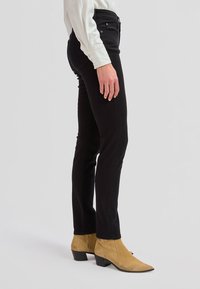 Osoba ubrana w czarne wąskie dżinsy, beżowe botki z ostrym noskiem i białą koszulę z długim rękawem, stojąca bokiem na neutralnym tle.