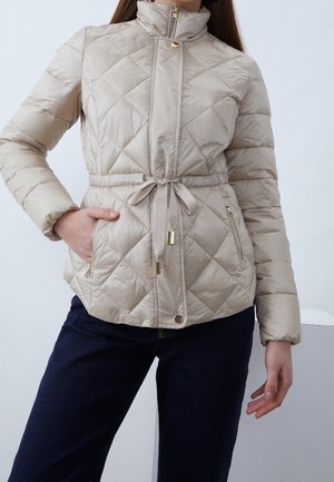 Vrouw met beige gewatteerd jack met voorrits, drukknopen, taillekoord en ritssluiting aan de zijzakken, gecombineerd met een donkere blauwe spijkerbroek.