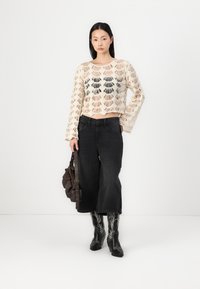 Sweater de crochê com bainha em forma de escamas em bege e preto, combinado com calças de ganga pretas de perna larga e botas de cowboy pretas bordadas. Bolsa castanha.