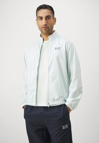 Corta-vento verde-mint com fecho éclair e textura em grelha, apresentando um colarinho alto e punhos elásticos, usado sobre uma blusa clara e calças escuras com marca.