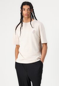 Beige kortærmet t-shirt med rund halsudskæring, prydet med blomsterbroderi i blå og pink på venstre bryst. Glat bomuldsstof.
