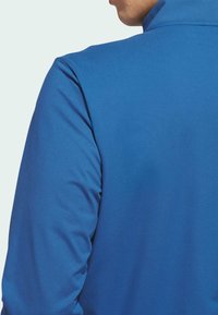 Plava sportska jakna s visokim ovratnikom, glatkom teksturom i pristajućim dizajnom. Ima bešavni prijelaz na ramenima i elegantan siluetu.