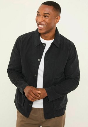 Black canvas jacket with a collared neckline, button closure, two front pockets, and a relaxed fit, worn over a white shirt.