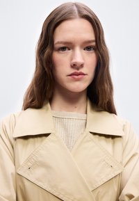Jeune femme aux longs cheveux bruns portant un trench-coat beige et un pull en maille crème, regardant directement l'appareil photo avec une expression neutre.
