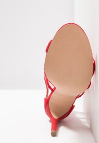 Červený vysoký podpatek se strukturovaným svrškem, otevřenou patou a tenkými kotníkovými pásky. Hladká béžová podešev s jemnými gripovými drážkami.