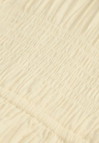 Krämfärgat tyg med en texturerad, sammanknuten design. Har parallella sömmar som skapar subtila vågor och ett mjukt, lätt utseende.