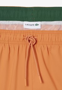 Oranje zwembroek met een elastische tailleband, een trekkoord vastgeknoopt, een witte mesh voering en een Lacoste-label met een groen krokodillenlogo.