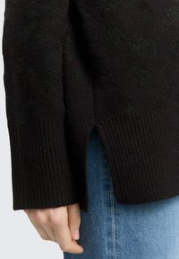 Zwarte gebreide trui met een getextureerd luipaardpatroon, geribbelde manchetten en zijsplitten. De stof lijkt zacht en warm. Gecombineerd met denim.