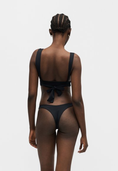 Femme aux cheveux tressés portant un bikini noir avec un nœud attaché dans le dos, tournée de dos sur un fond gris uni.