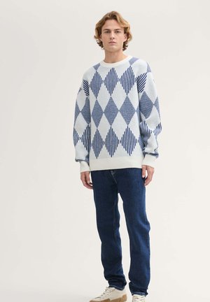 Suéter de patrón de diamante blanco y azul con cuello redondo, mangas largas y puños de canalé, combinado con jeans azul oscuro y zapatillas color beige.