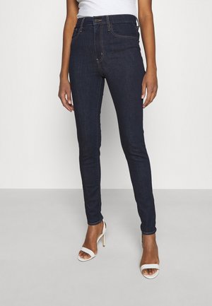 Femme portant un jean skinny bleu foncé et des sandales blanches à talons hauts, debout devant un fond clair uni.