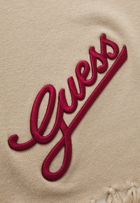 Tissu beige avec un logo "Guess" en filigrane burgonde en écriture cursive, avec un bord effiloché en bas.
