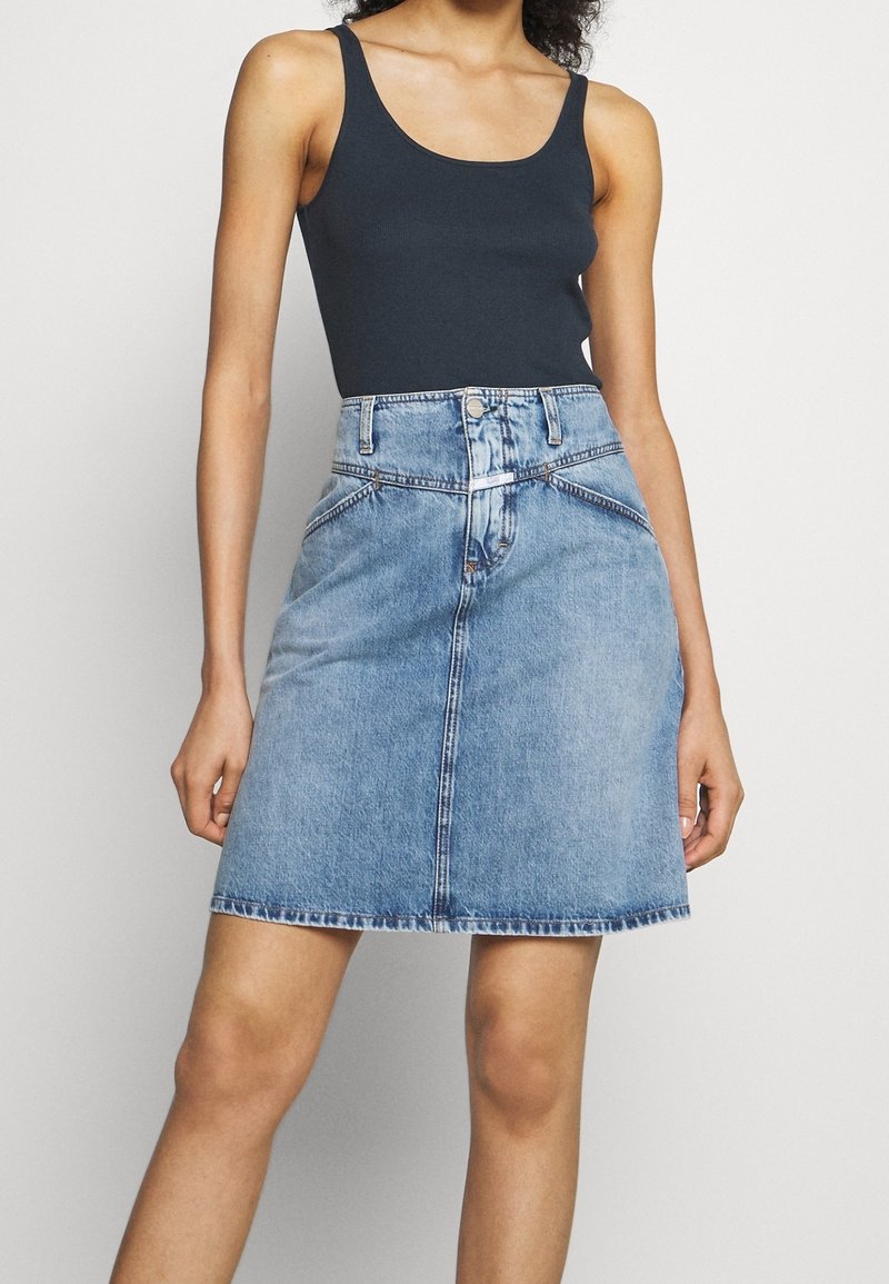 Femme portant un haut noir sans manches et une jupe en jean bleue taille haute, debout devant un fond uni.