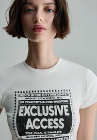 Fehér pamut póló, amely grafikus nyomattal rendelkezik, fekete szöveggel és ezüst szegecsekkel a dizájn körül, amely így szól: "Kizárólagos hozzáférés minden színpadhoz".