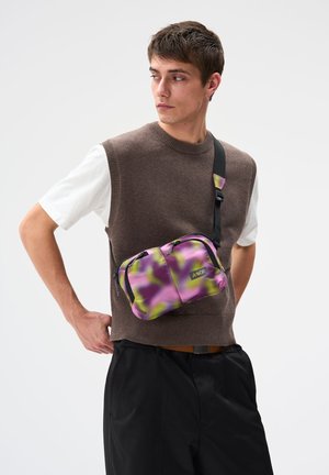 Junger Mann, der einen braunen Ärmellosen Pullover, ein weißes T-Shirt, schwarze Hosen und eine lila-grüne Tarn-Umhängetasche trägt und nach links schaut.