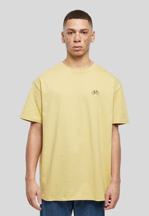 Jeune homme aux cheveux courts et aux taches de rousseur, portant un t-shirt jaune pâle avec un petit motif de vélo sur la poitrine, se tenant devant un fond gris uni.