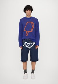 Sweater azul com um design de picolé laranja, combinado com calções de ganga que apresentam uma gráfica de um cão de desenhos animados em branco. Ténis brancos e meias cinzentas.