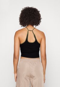 En person med lockigt hår bär en svart crop top med grön kant och beige byxor, sedd från ryggen mot en neutral bakgrund.