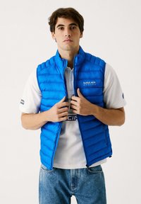 Blå quilted vest med let polstring, lynlås foran, og Red Bull Racing-logo på brystet, lagd over en hvid polo skjorte.