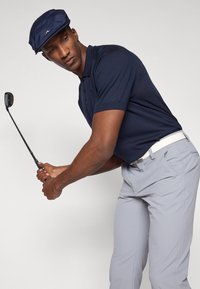 Marine polo-shirt med knappet krage, lysegrå bukser og en marin cap. Personen holder en golfklubbe og viser en svingstilling.