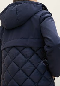 Námorní pufr jacket s prošívanou texturou, obsahující kapuci a hladké ramenní části. Design zahrnuje kontrast mezi matnými a lesklými povrchy.