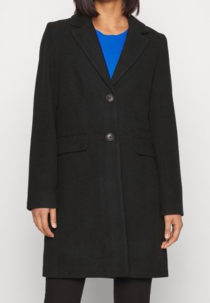 Person trägt einen schwarzen Mantel mittlerer Länge mit zwei Knöpfen und Klappentaschen über einem blauen Oberteil, steht vor einem schlichten Hintergrund.