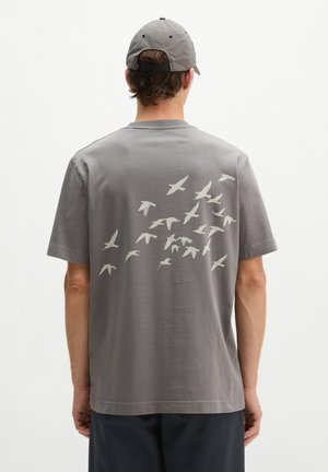 Mann trägt graues T-Shirt und Kappe mit weißen Vogelsilhouetten, die diagonal über den Rücken des Shirts fliegen.