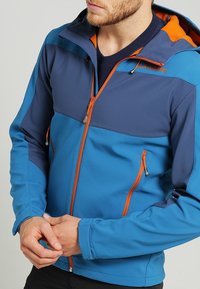 Blauwe softshelljack met een color-block ontwerp en marineblauwe en oranje accenten, voorkant met ritssluiting en zijzakken met rits.