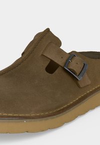 Καφέ παπούτσι από καστόρι τύπου slip-on με ρυθμιζόμενο λουράκι και μπεζ καουτσούκ σόλα, μερικώς ορατό σε ανοιχτό φόντο.